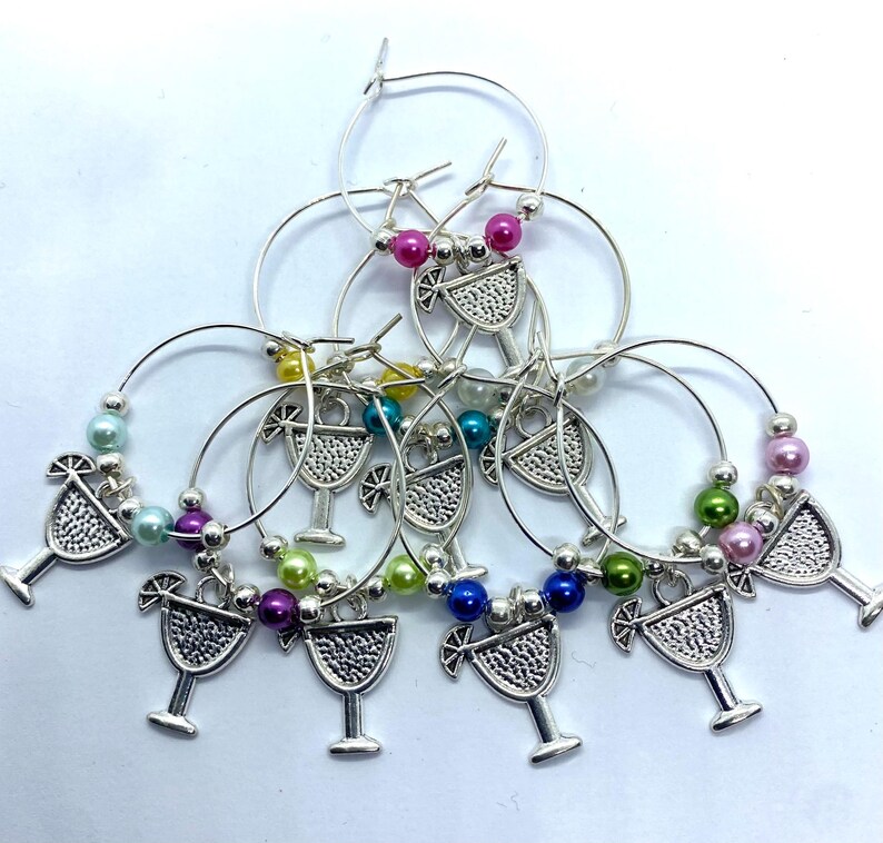 May include: A collection of silver-toned hoop wine glass charms, each featuring a textured cocktail glass charm and accented with colorful round beads in shades of pink, blue, green, yellow, and purple, arranged in a layered circular pattern.