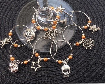May include: Set of six Halloween-themed wine glass charms. Each charm features a different Halloween icon, including a spider, a pumpkin, a bat, a witch, a skull, and a spiderweb. The charms are made of silver metal and have orange beads.