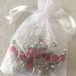 May include: A white organza gift bag tied with a ribbon, containing several silver wine glass charms. Each charm has red and green beads and a snowflake charm.