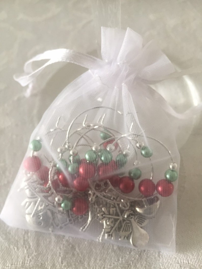 May include: A white organza gift bag tied with a ribbon, containing several silver wine glass charms. Each charm has red and green beads and a snowflake charm.