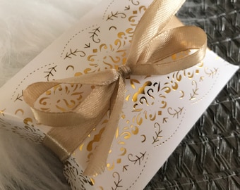 May include: A white favor box with a gold, intricate, laser-cut design and a gold satin ribbon tied in a bow.