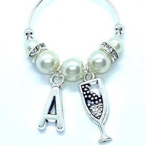 May include: A silver hoop wine glass charm featuring three white faux pearls, two silver-toned spacers with embedded clear crystals, a silver-toned letter A charm, and a silver-toned champagne flute charm with black detailing on the glass.