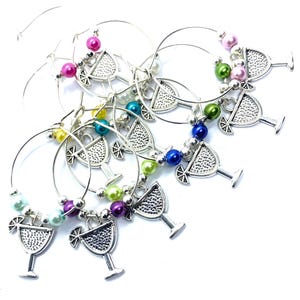 May include: A set of silver-toned metal wine glass charms featuring a cocktail glass pendant with a citrus slice garnish. Each circular wire hoop is decorated with a variety of colorful, glossy round beads in shades of pink, green, blue, and purple.