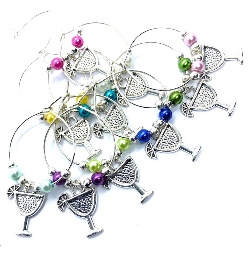 May include: A set of silver-toned metal wine glass charms featuring a cocktail glass pendant with a citrus slice garnish. Each circular wire hoop is decorated with a variety of colorful, glossy round beads in shades of pink, green, blue, and purple.
