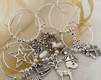 May include: Silver-toned Christmas-themed wine charms. Each charm features a different holiday design, including a star, snowflake, snowman, Christmas tree, and gift. The charms are attached to silver hoops with faux pearl accents.