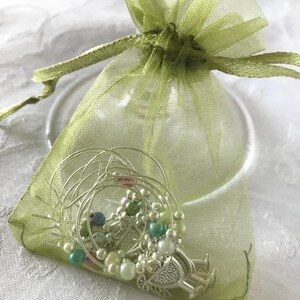 May include: A sheer light green organza drawstring pouch containing several silver-toned wire wine glass charms. The charms feature small round beads in shades of white, teal, and green, along with decorative metal accents shaped like wine glasses.