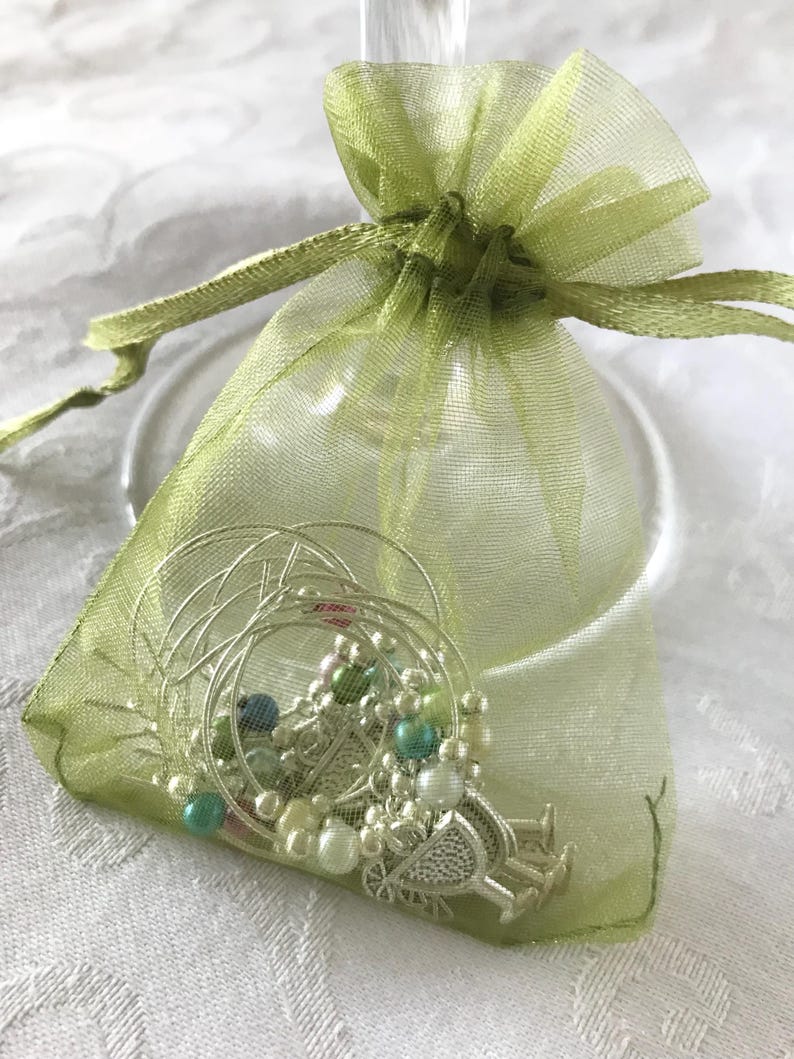 May include: A sheer light green organza drawstring pouch containing several silver-toned wire wine glass charms. The charms feature small round beads in shades of white, teal, and green, along with decorative metal accents shaped like wine glasses.
