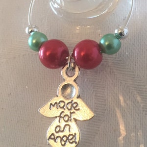 May include: A wine glass charm with a silver angel pendant that reads "Made for an Angel." The charm features red and green beads, and a silver bead. The charm is designed to hang on a wine glass.