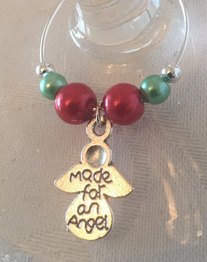 May include: A wine glass charm with a silver angel pendant that reads "Made for an Angel." The charm features red and green beads, and a silver bead. The charm is designed to hang on a wine glass.
