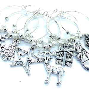 May include: A set of silver-toned metal wine glass charms featuring holiday-themed pendants including a snowflake, Christmas tree, star, reindeer, gift box, snowman, and stocking, accented with small white beads on circular wire hoops.
