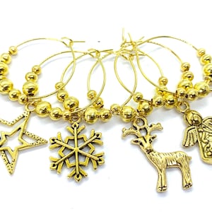 May include: A set of gold-toned metal wine glass charms featuring various holiday-themed pendants including snowflakes, a star, a reindeer, an angel, and a figure, all attached to circular hoops decorated with small gold beads.