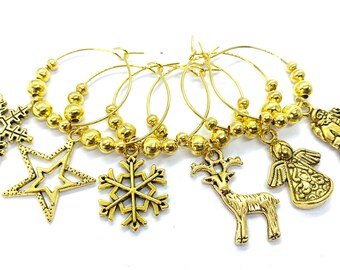 May include: A set of gold-toned metal wine glass charms featuring various holiday-themed pendants including snowflakes, a star, a reindeer, an angel, and a figure, all attached to circular hoops decorated with small gold beads.