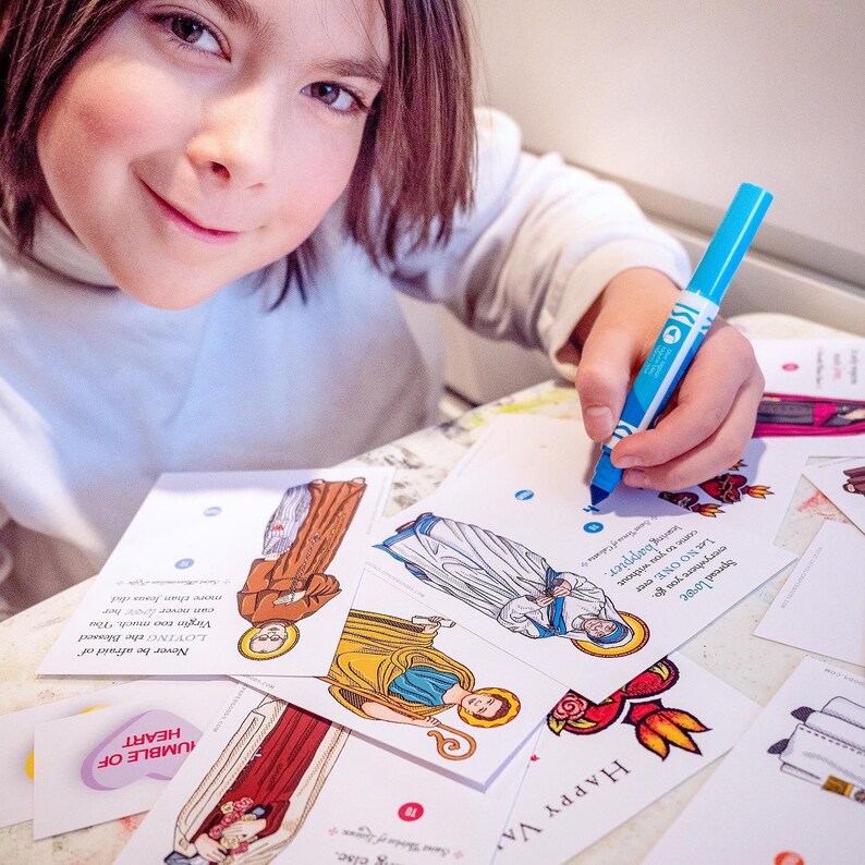 Puede incluir: Un ni&ntilde;o colorea un conjunto de tarjetas para colorear de tem&aacute;tica religiosa con un marcador azul. Las tarjetas presentan ilustraciones de santos y figuras religiosas. El texto en las tarjetas incluye frases como "Difunde el amor" y "Nunca tengas miedo de amar demasiado a la Virgen Mar&iacute;a".