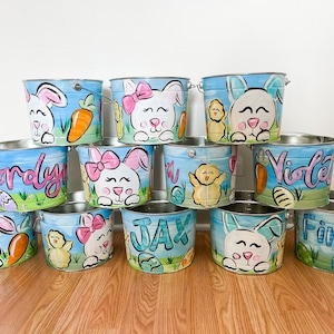 May include: A set of ten blue and white painted metal buckets with bunny and chick designs. Each bucket has a different name painted on it, including Jordyn, Jax, Finley, and Violet.