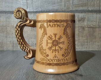 Può includere: Un boccale di birra in legno chiaro con manico e corpo intagliati. Il boccale è inciso con il nome "JAMES" sopra un simbolo vichingo e la parola "GROOMSMAN" sotto. Il manico ha un design a voluta decorativa.