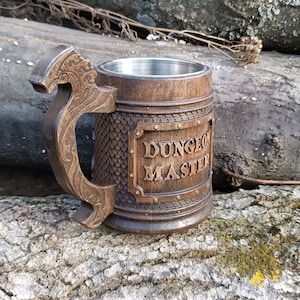 Puede incluir: Una jarra de cerveza de madera con un interior de acero inoxidable. La jarra tiene un asa en forma de dragón y un diseño tallado de escamas. La parte delantera de la jarra tiene una placa de metal con las palabras "Dungeon Master" grabadas en ella.