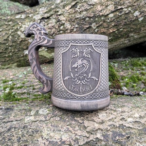 Könnte beinhalten: Ein brauner Bierkrug aus Holz mit einem Drachendesign. Der Krug weist aufwendige Schnitzereien auf, darunter ein Drachenwappen und den Namen "Michael". Der Griff ist als Drachenkopf geformt. Der Krug liegt auf einem Baumstamm.