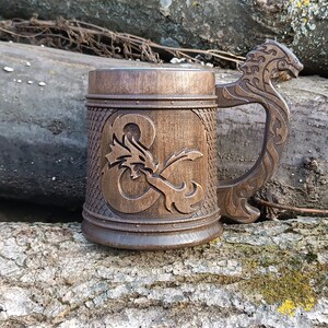 May include: A wooden beer mug with a carved dragon and the Dungeons & Dragons logo. The handle is intricately carved with a dragon head design. The mug is brown and sits on a log.