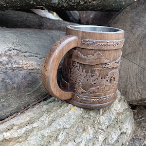 Pode incluir: Caneca de madeira entalhada &agrave; m&atilde;o com uma cena detalhada da floresta. A caneca apresenta uma al&ccedil;a grande e curva e um interior de a&ccedil;o inoxid&aacute;vel. A madeira marrom escura contrasta com o interior prateado.
