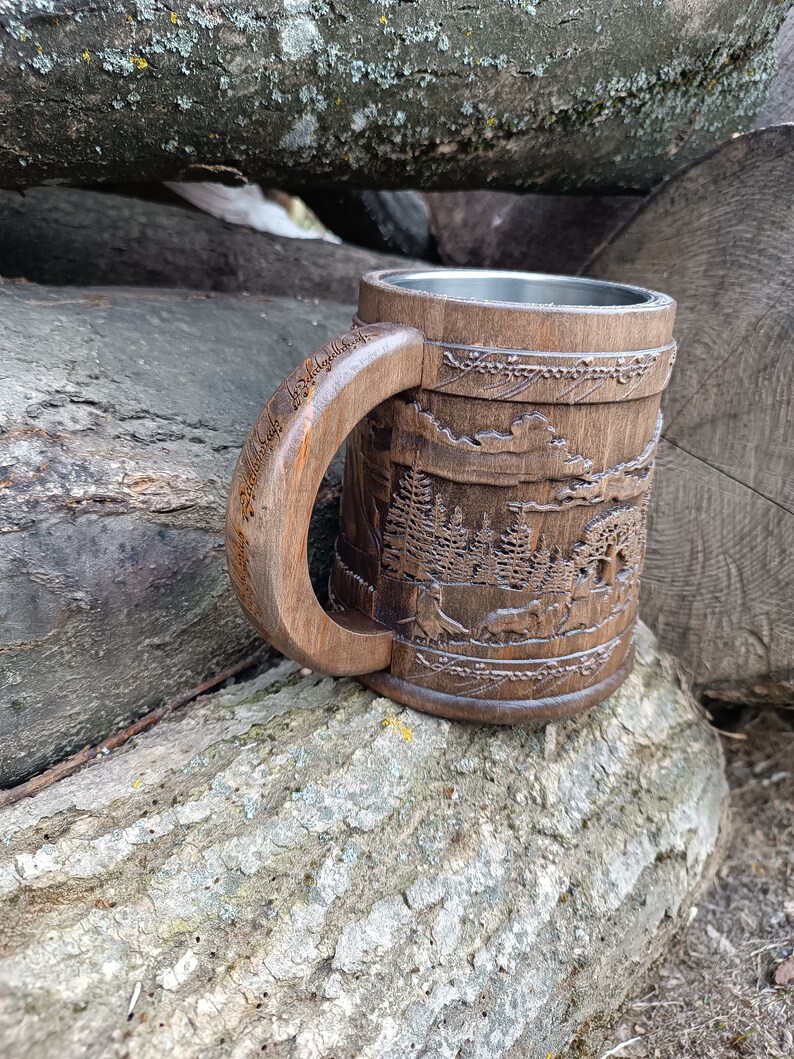 Pode incluir: Caneca de madeira entalhada &agrave; m&atilde;o com uma cena detalhada da floresta. A caneca apresenta uma al&ccedil;a grande e curva e um interior de a&ccedil;o inoxid&aacute;vel. A madeira marrom escura contrasta com o interior prateado.