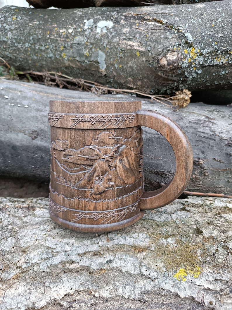 Pode incluir: Uma caneca de madeira castanha escura com uma al&ccedil;a grande. A caneca apresenta imagens esculpidas de uma paisagem montanhosa e texto na parte superior e inferior. A caneca est&aacute; sobre uma superf&iacute;cie de madeira texturizada.