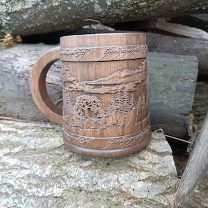 Pode incluir: Caneca de madeira castanha com um desenho de paisagem entalhado e texto. A caneca tem uma pega grande. O desenho entalhado inclui &aacute;rvores, montanhas e texto ao redor da parte superior. A caneca est&aacute; sobre uma superf&iacute;cie cinza texturizada.