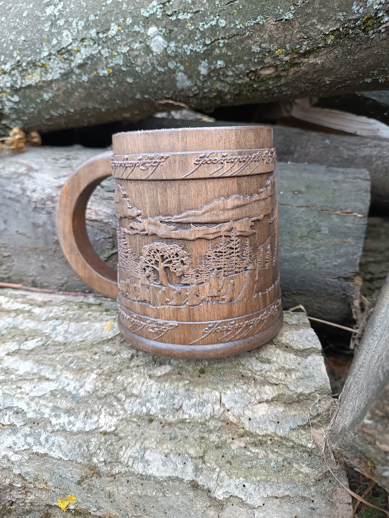 Pode incluir: Caneca de madeira castanha com um desenho de paisagem entalhado e texto. A caneca tem uma pega grande. O desenho entalhado inclui &aacute;rvores, montanhas e texto ao redor da parte superior. A caneca est&aacute; sobre uma superf&iacute;cie cinza texturizada.