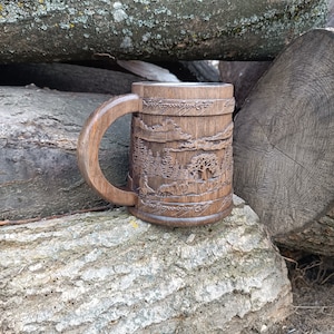 Pode incluir: Uma caneca de madeira entalhada &agrave; m&atilde;o com um design de paisagem. A caneca apresenta uma al&ccedil;a grande e detalhes intrincados de &aacute;rvores, montanhas e vida selvagem. A caneca marrom escura est&aacute; sobre um fundo de toras de madeira.