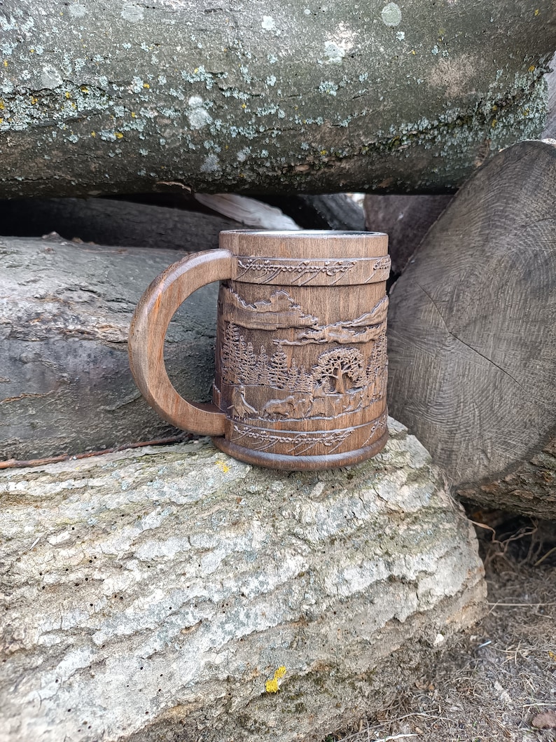 Pode incluir: Uma caneca de madeira entalhada &agrave; m&atilde;o com um design de paisagem. A caneca apresenta uma al&ccedil;a grande e detalhes intrincados de &aacute;rvores, montanhas e vida selvagem. A caneca marrom escura est&aacute; sobre um fundo de toras de madeira.