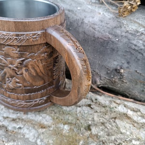 Pode incluir: Caneca de madeira castanha com interior em a&ccedil;o inoxid&aacute;vel. O exterior apresenta imagens e texto esculpidos, com uma pega que tamb&eacute;m apresenta texto gravado. A caneca tem um aspeto artesanal e um design r&uacute;stico.