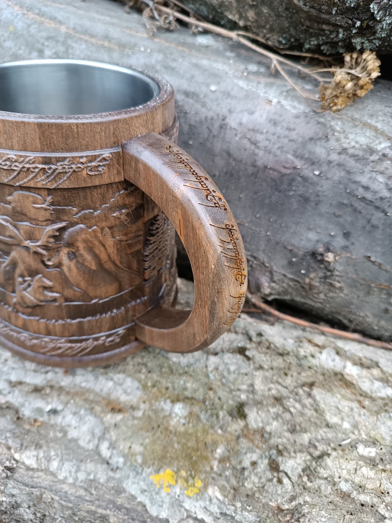 Pode incluir: Caneca de madeira castanha com interior em a&ccedil;o inoxid&aacute;vel. O exterior apresenta imagens e texto esculpidos, com uma pega que tamb&eacute;m apresenta texto gravado. A caneca tem um aspeto artesanal e um design r&uacute;stico.