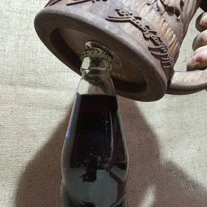 Pode incluir: Um caneco de cerveja de madeira entalhada castanha com um abridor de garrafas embutido na base. Uma garrafa de vidro transparente cheia de um l&iacute;quido escuro est&aacute; sendo aberta. O caneco tem uma al&ccedil;a e entalhes decorativos.