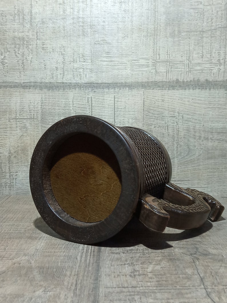 Pu&ograve; includere: Una tazza di legno marrone scuro con un manico a forma di drago scolpito. La tazza ha una parte superiore rotonda aperta e un motivo intrecciato testurizzato attorno al corpo.