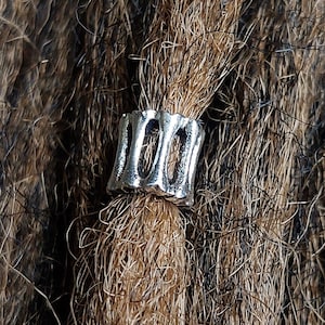 May include: A silver-colored dreadlock bead with a textured, open design. The bead is cylindrical and wraps around a section of brown and black dreadlocks, adding a decorative element to the hair.