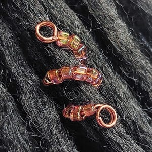 May include: Close-up of three copper-colored dreadlock beads. Each bead features a spiral design with a loop at the end. The beads are adorned with small, iridescent, rectangular beads in shades of orange, yellow, and pink, creating a shimmering effect.
