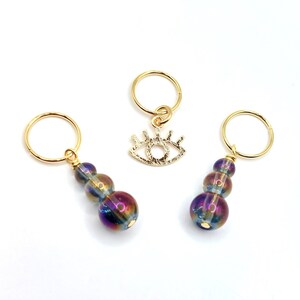 May include: Three gold stitch markers with iridescent glass beads. One marker features a gold eye charm.