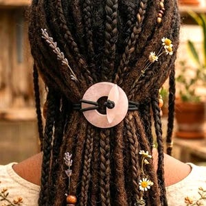 May include: A person's dark brown dreadlocks are styled with braids and adorned with small white flowers, lavender sprigs, and wooden beads. A light pink stone hair tie secures the hair. The person wears a beige shirt with floral embroidery.
