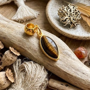 May include: A gold pendant with an oval tiger's eye stone is displayed on a piece of driftwood. The pendant is surrounded by other natural elements, including crystals, dried flowers, and twine. The pendant's stone has brown and gold tones.