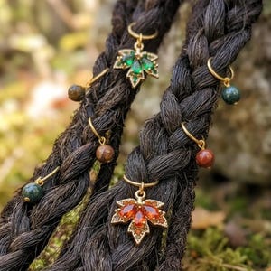 May include: Close-up of two dark braids adorned with gold-colored hair jewelry. The jewelry includes leaf-shaped charms with green and orange gemstones, and small beaded accents. The background is blurred, with green moss and foliage.