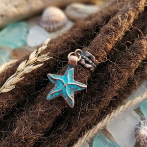 May include: A copper-colored starfish charm with turquoise accents is attached to a brown dreadlock. The charm is part of a hair accessory. The background includes seashells, sea glass, and a piece of driftwood.