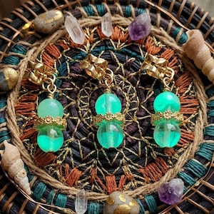 May include: Three pairs of earrings with a gold-colored floral design at the top and a double-beaded, glowing green orb. The earrings are displayed on a woven basket with crystals, stones, and seashells.