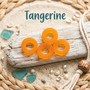 May include: Four circular, translucent orange beads with a matte finish are arranged on a piece of weathered driftwood. The word Tangerine is printed above the beads. The driftwood rests on light-colored sand surrounded by small sea shells.