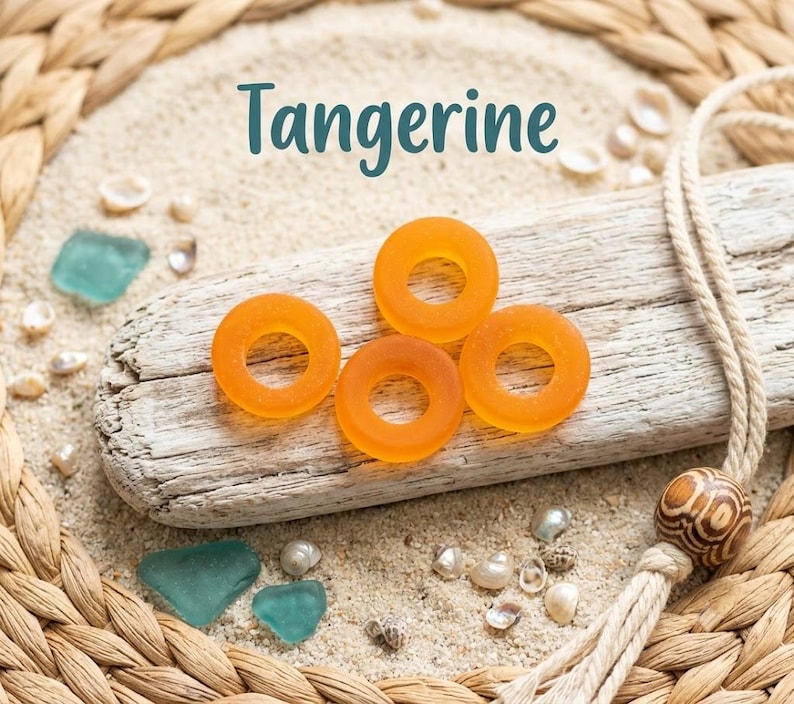 May include: Four circular, translucent orange beads with a matte finish are arranged on a piece of weathered driftwood. The word Tangerine is printed above the beads. The driftwood rests on light-colored sand surrounded by small sea shells.