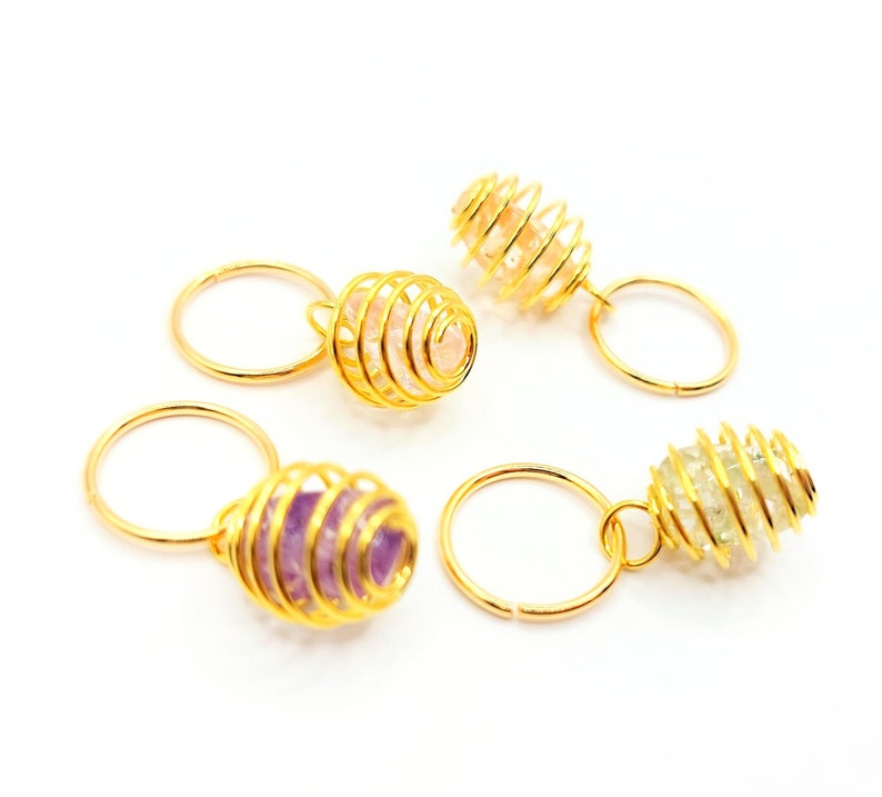 May include: Four gold wire wrapped crystal cages with different colored crystals. The crystals are pink, purple, and clear. The cages are attached to gold rings.