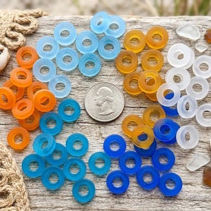 May include: A collection of circular, translucent glass beads in shades of blue, orange, and white, arranged around a United States quarter coin for scale. The beads feature a smooth, matte finish and a central hole, accompanied by two small seashells.