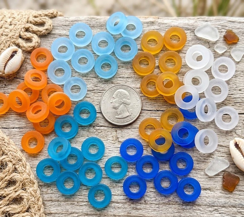 May include: A collection of circular, translucent glass beads in shades of blue, orange, and white, arranged around a United States quarter coin for scale. The beads feature a smooth, matte finish and a central hole, accompanied by two small seashells.