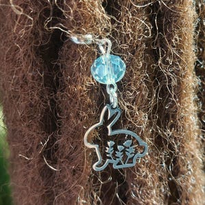 May include: A silver charm with a cutout of a rabbit and flowers hanging from a silver chain with a light blue crystal bead.