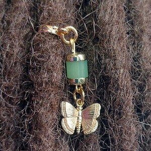 May include: Gold-toned hair accessory featuring a light green cylindrical bead and a butterfly charm. The butterfly has detailed wing patterns. The accessory is attached to brown hair.