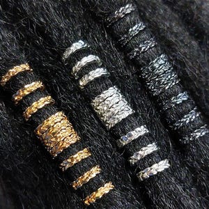 May include: Close-up of several dreadlock beads in gold, silver, and gunmetal colors. The beads have various designs, including leaf and textured patterns, and are set against a backdrop of dark hair.
