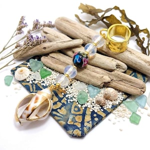 May include: A gold ring and a seashell pendant with a beaded strand, arranged with driftwood, sea glass, and small seashells on a patterned blue fabric. The beaded strand features clear and colorful glass beads.